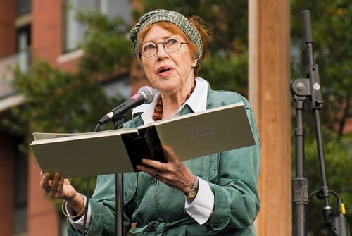 Kathleen Fraser wearing wire-rimmed glasses, looking upward and to the right, holding a large book at a podium