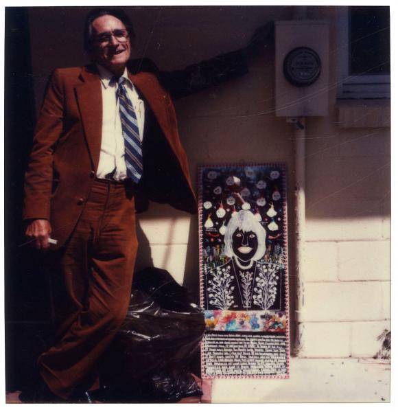 Howard Finster in a brown suit leaning against a wall next to a sidewalk advertisement