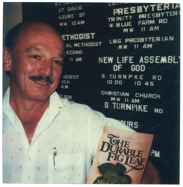 Ronald H. Bayes standing in front of a bulletin board holding a copy of 'The Durable Fig Leaf'