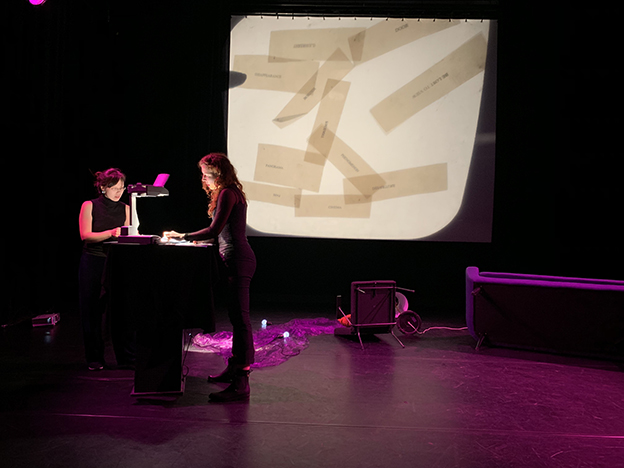 two women standing on-stage at an overhead projector; projector image on wall behind has translucent strips with text scattered on it 