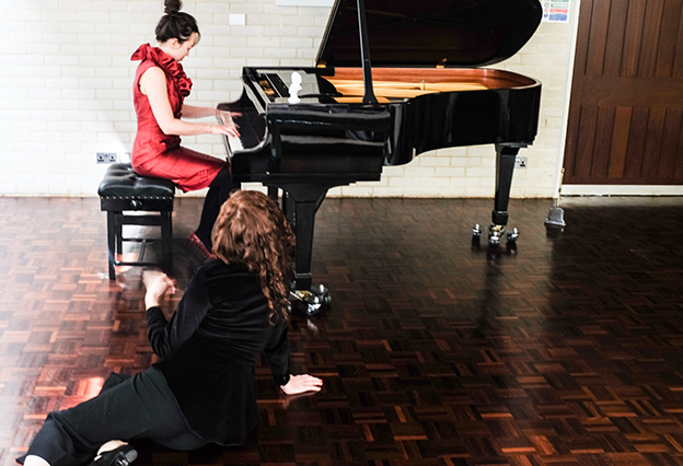 woman seated and playing piano with another woman facing away from the camera seated on the floor