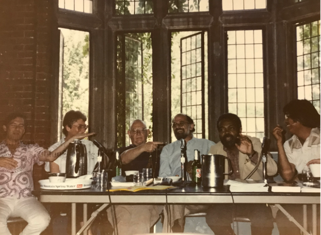 People surround Amiri Baraka who speaks at a table in front of an open window. 