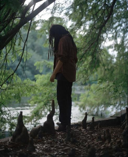 Taylor Johnson with side/back to camera and long locs peering over the edge of a dropoff over water, surrounded by knobby roots that poke straight up near the edge