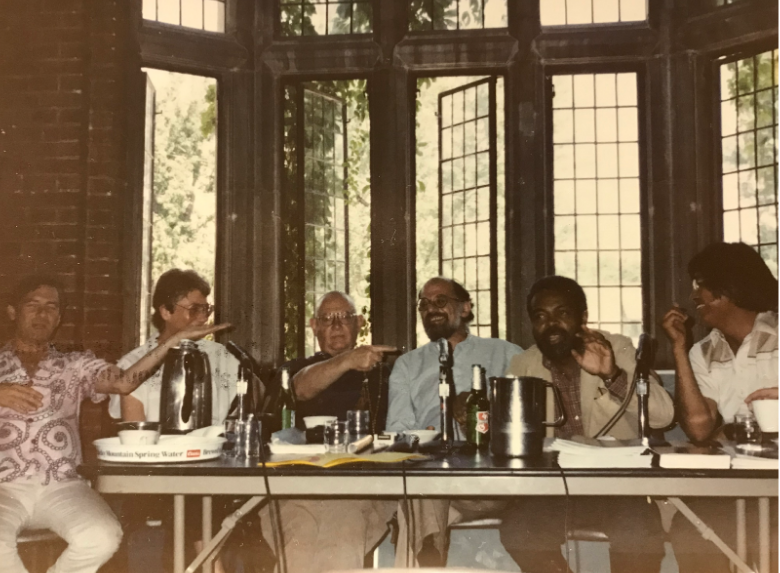 Unattributed photo from the Naropa Archives. Photographed in the archive by Jaime Groetsema. People surround Amiri Baraka who speaks at a table in front of an open window.
