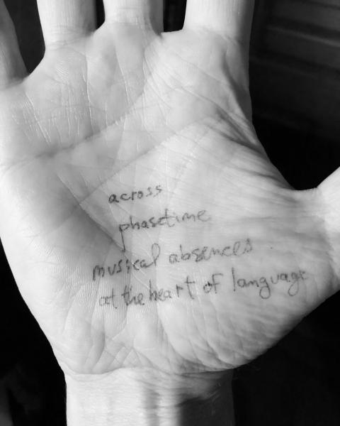 Person's palm with words written in pen on it: "across / phasetime / musical absences / at the heart of language."
