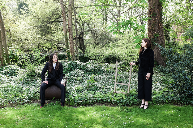 two women in suits in front of a wooded area, one seated, one standing, spaced six feet apart